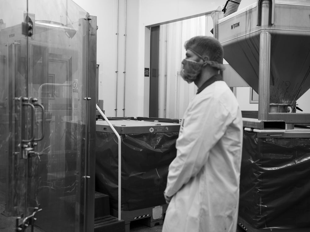 A man in a lab coat and hairnet on the factory floor, beside bulk powder bins